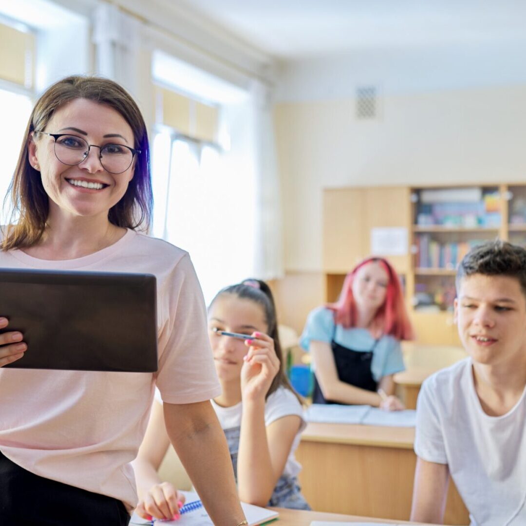 Portrait of female teacher at school at lesson, smiling middle-aged woman in glasses with digital tablet looking at camera, classroom with teenage students at desk background